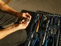 Mains féminines avec ongles blancs, manipulant une clé à cliquet. Une boîte à outils bleue est ouverte à côté, remplie d'outils. Une scène de travaux sans accord.