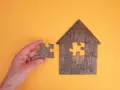 Main d'une personne tenant une pièce de puzzle pour compléter une maison en bois. Une métaphore pour la donation-partage indivision.