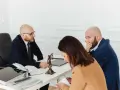 Trois personnes à une table, dont un homme en costume parlant à un autre homme qui se couvre la bouche.