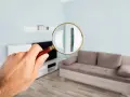 Une loupe examine un salon moderne. On voit un canapé beige, une étagère murale et une télévision. Le mur est bleu pâle, et le parquet est clair. Le plu bordeaux est peut-être caché quelque part.