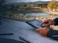 Un homme consulte une tablette et une carte sur le capot d'une voiture, avec un paysage de campagne et un étang en arrière-plan, rappelant une exploration près de **Plu Aix-en-Provence**.