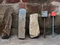 Quatre bornes, dont une borne cadastre en pierre, alignées devant un mur de pierres anciennes.