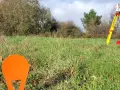 Un géomètre utilise un théodolite sur un trépied jaune pour relever des points sur un terrain vague, peut-être pour le cadastre de Sarreguemines.