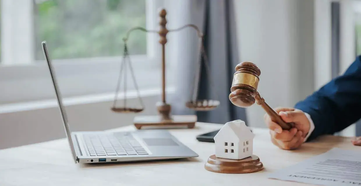 Marteau de juge frappant une maison miniature, symbolisant la loi Carrez pour une maison individuelle. Balance de la justice et ordinateur en arrière-plan.