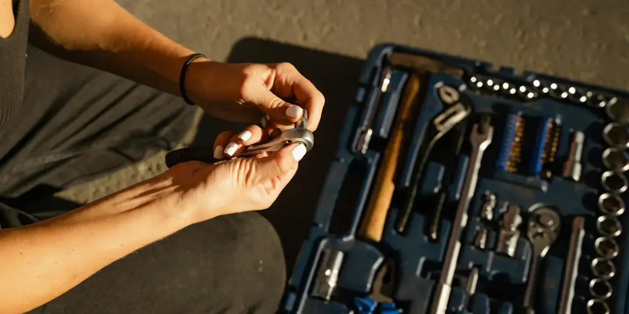 Mains féminines avec ongles blancs, manipulant une clé à cliquet. Une boîte à outils bleue est ouverte à côté, remplie d'outils. Une scène de travaux sans accord.