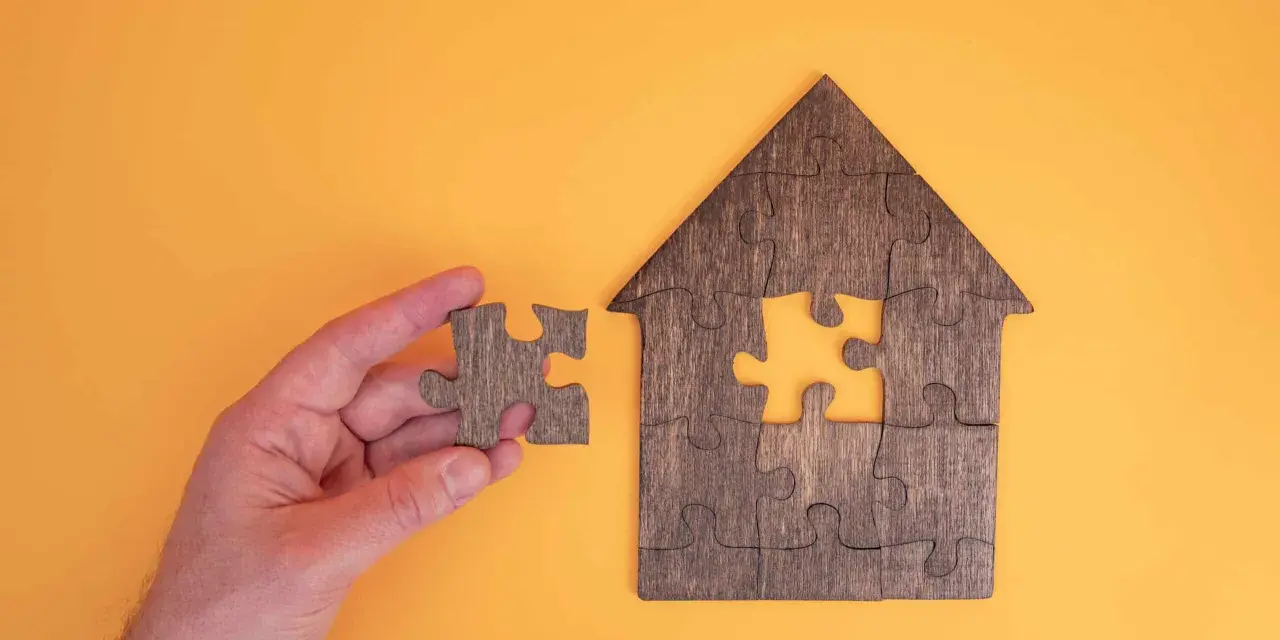 Main d'une personne tenant une pièce de puzzle pour compléter une maison en bois. Une métaphore pour la donation-partage indivision.