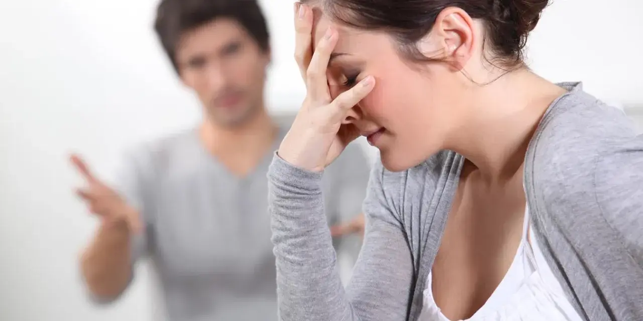 Couple en conflit, femme se tenant le front, homme parlant au fond.