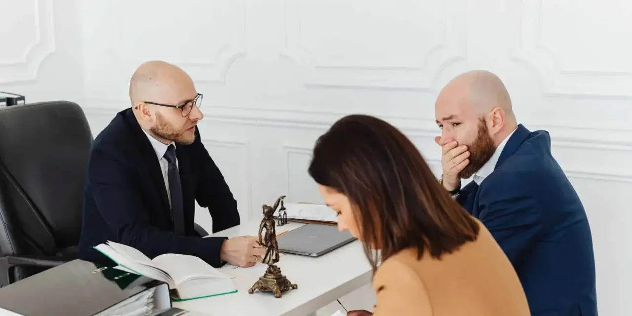 Trois personnes à une table, dont un homme en costume parlant à un autre homme qui se couvre la bouche.