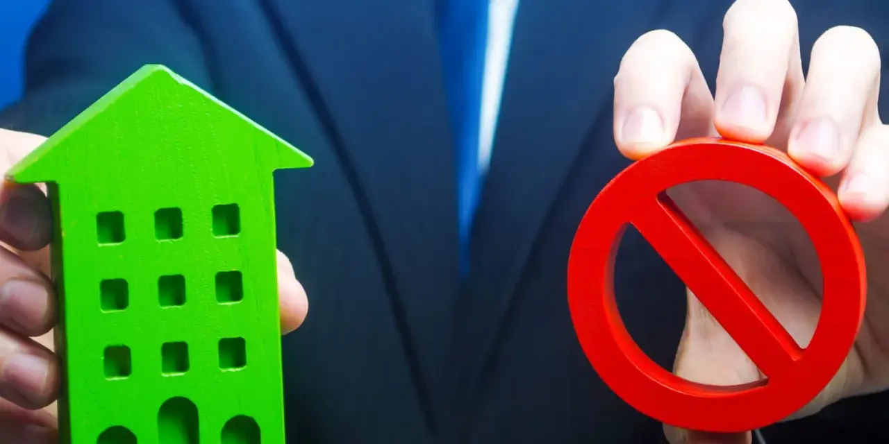 Un homme en costume tient une maison verte et un panneau d'interdiction rouge, symbolisant l'opposition au prix de vente chez le notaire.