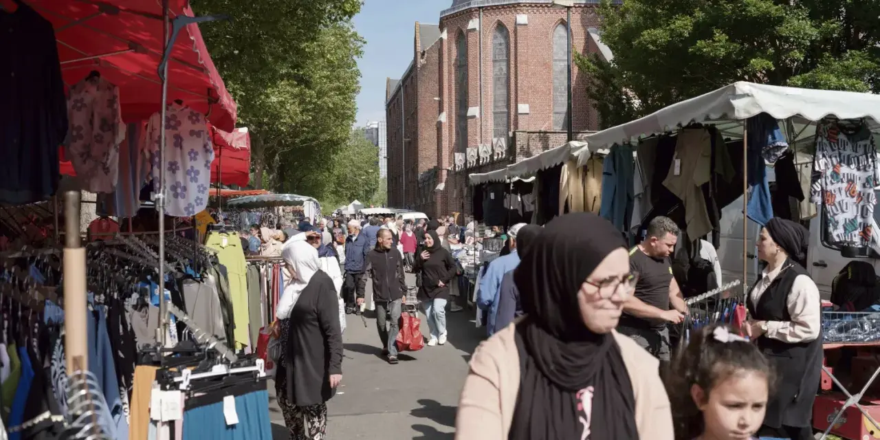 Ambiance de marché animé à **Plu Roubaix**, avec des étals de vêtements colorés et des passants sous le soleil.