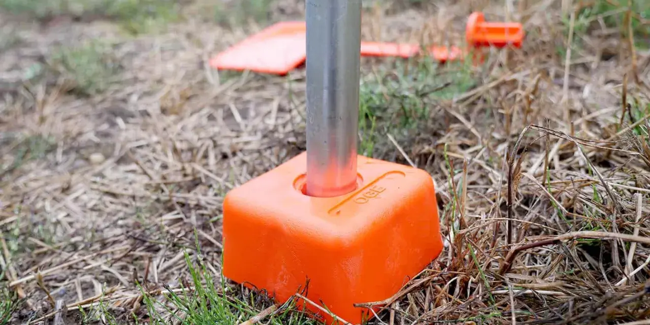 Borne orange avec un tube métallique planté dans le sol. D'autres bornes sont visibles en arrière-plan, indiquant le bornage d'un terrain déjà borné.