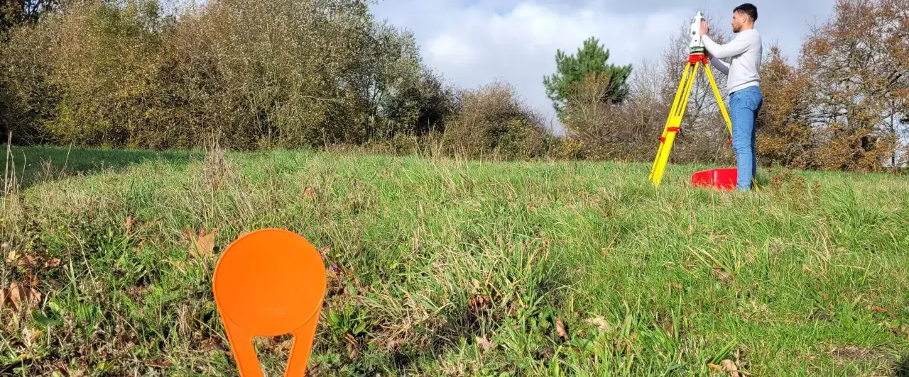Un géomètre utilise un théodolite sur un trépied jaune pour relever des points sur un terrain vague, peut-être pour le cadastre de Sarreguemines.
