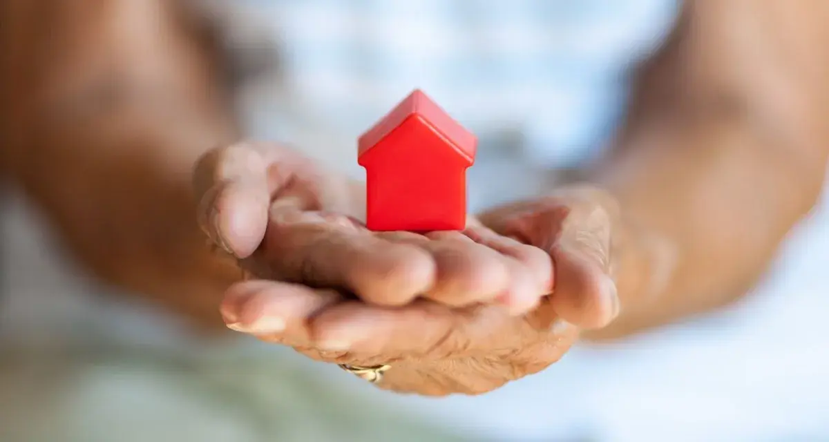 Mains âgées tenant une petite maison rouge. Une image symbolisant la "cosaisine définition" : le foyer, la sécurité, le bien-être.