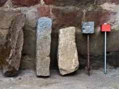 Quatre bornes, dont une borne cadastre en pierre, alignées devant un mur de pierres anciennes.