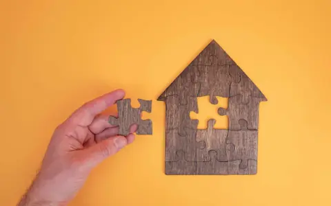 Main d'une personne tenant une pièce de puzzle pour compléter une maison en bois. Une métaphore pour la donation-partage indivision.