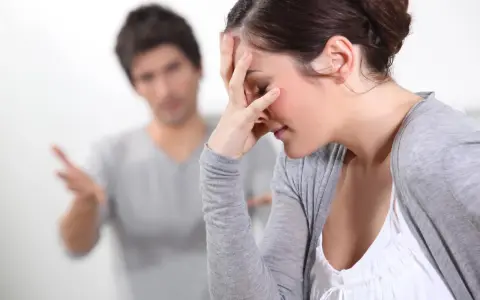 Couple en conflit, femme se tenant le front, homme parlant au fond.