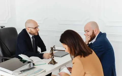 Trois personnes à une table, dont un homme en costume parlant à un autre homme qui se couvre la bouche.