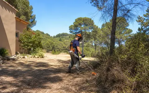 Un homme équipé pour le débroussaillement travaille près d'une maison, dans un paysage boisé. La jurisprudence débroussaillement est essentielle pour la prévention des incendies.