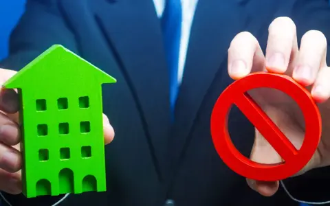 Un homme en costume tient une maison verte et un panneau d'interdiction rouge, symbolisant l'opposition au prix de vente chez le notaire.