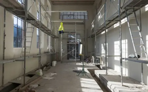 Chantier intérieur avec échafaudages, un gilet jaune suspendu, des matériaux de construction et des ouvertures. Un **daact non conforme** pourrait être la raison de cette scène.