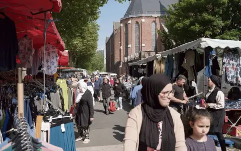 Ambiance de marché animé à **Plu Roubaix**, avec des étals de vêtements colorés et des passants sous le soleil.