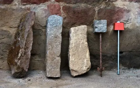 Quatre bornes, dont une borne cadastre en pierre, alignées devant un mur de pierres anciennes.