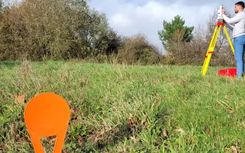 Un géomètre utilise un théodolite sur un trépied jaune pour relever des points sur un terrain vague, peut-être pour le cadastre de Sarreguemines.