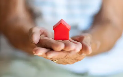 Mains âgées tenant une petite maison rouge. Une image symbolisant la "cosaisine définition" : le foyer, la sécurité, le bien-être.