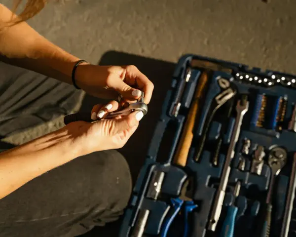 Mains féminines avec ongles blancs, manipulant une clé à cliquet. Une boîte à outils bleue est ouverte à côté, remplie d'outils. Une scène de travaux sans accord.
