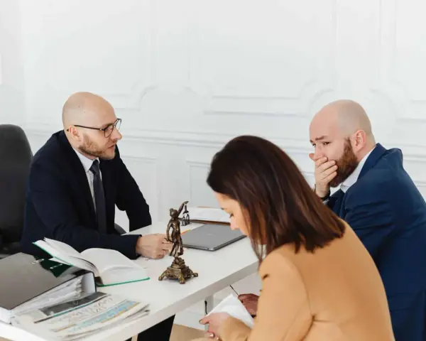 Trois personnes à une table, dont un homme en costume parlant à un autre homme qui se couvre la bouche.