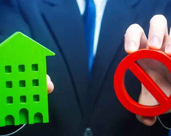 Un homme en costume tient une maison verte et un panneau d'interdiction rouge, symbolisant l'opposition au prix de vente chez le notaire.