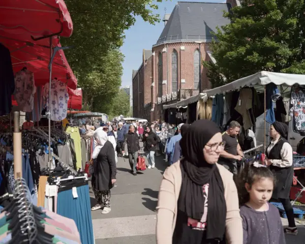 Ambiance de marché animé à **Plu Roubaix**, avec des étals de vêtements colorés et des passants sous le soleil.