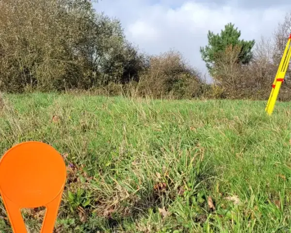 Un géomètre utilise un théodolite sur un trépied jaune pour relever des points sur un terrain vague, peut-être pour le cadastre de Sarreguemines.