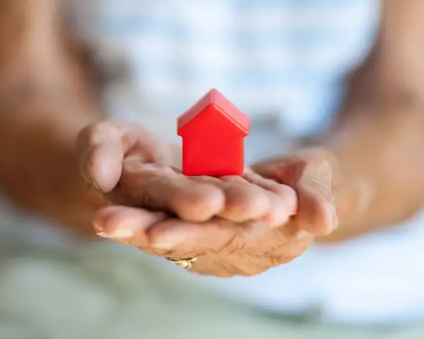 Mains âgées tenant une petite maison rouge. Une image symbolisant la "cosaisine définition" : le foyer, la sécurité, le bien-être.