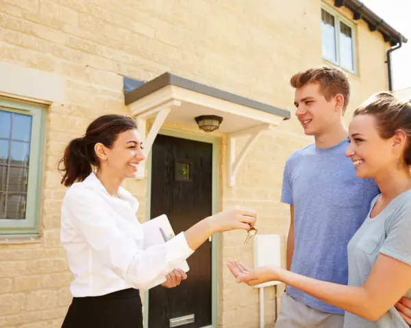 Une femme remet des clés à un jeune couple devant leur nouvelle maison. Le tableau droit de préemption locataire est respecté.