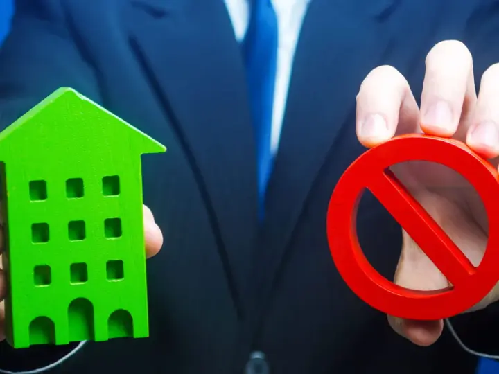 Un homme en costume tient une maison verte et un panneau d'interdiction rouge, symbolisant l'opposition au prix de vente chez le notaire.