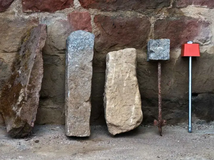 Quatre bornes, dont une borne cadastre en pierre, alignées devant un mur de pierres anciennes.