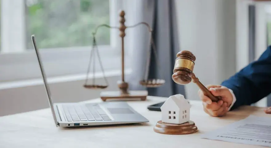 Marteau de juge frappant une maison miniature, symbolisant la loi Carrez pour une maison individuelle. Balance de la justice et ordinateur en arrière-plan.