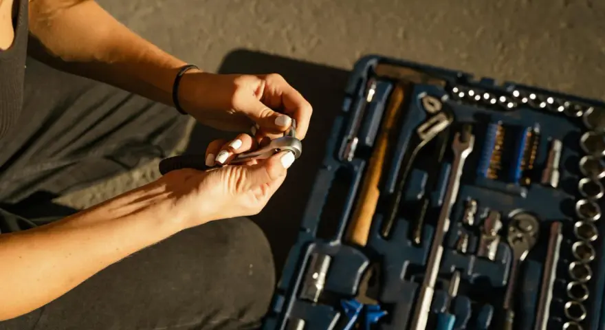 Mains féminines avec ongles blancs, manipulant une clé à cliquet. Une boîte à outils bleue est ouverte à côté, remplie d'outils. Une scène de travaux sans accord.