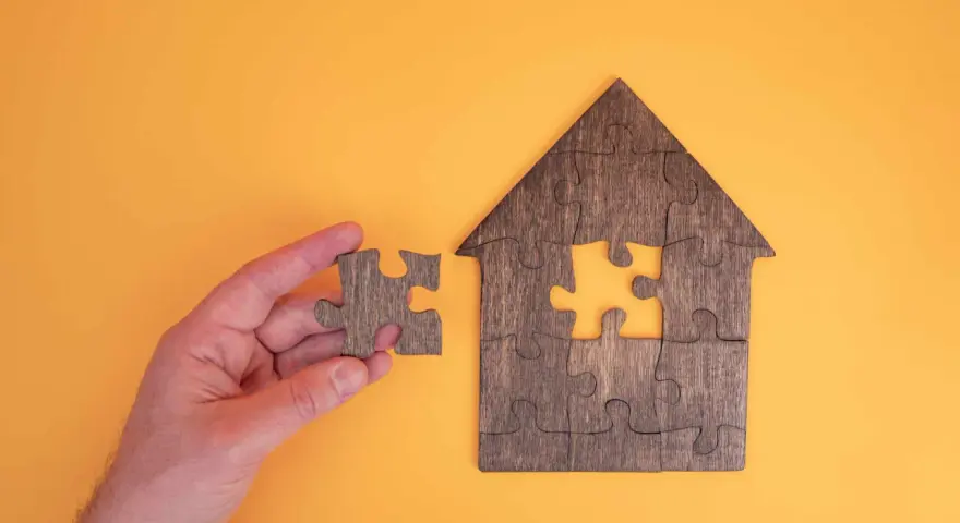 Main d'une personne tenant une pièce de puzzle pour compléter une maison en bois. Une métaphore pour la donation-partage indivision.