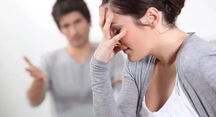 Couple en conflit, femme se tenant le front, homme parlant au fond.