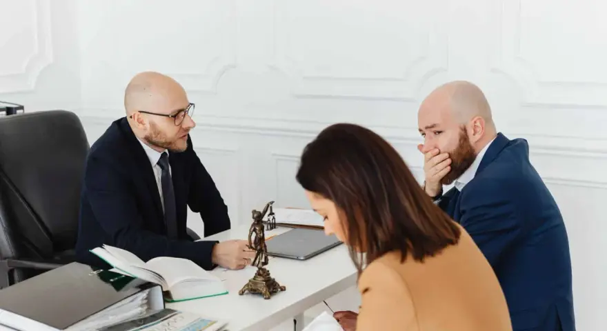 Trois personnes à une table, dont un homme en costume parlant à un autre homme qui se couvre la bouche.