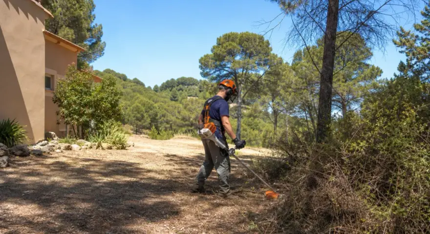 Un homme équipé pour le débroussaillement travaille près d'une maison, dans un paysage boisé. La jurisprudence débroussaillement est essentielle pour la prévention des incendies.