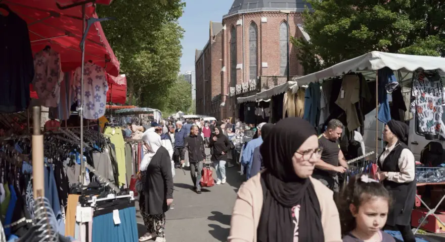 Ambiance de marché animé à **Plu Roubaix**, avec des étals de vêtements colorés et des passants sous le soleil.