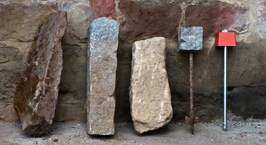 Quatre bornes, dont une borne cadastre en pierre, alignées devant un mur de pierres anciennes.