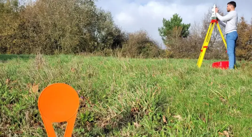 Un géomètre utilise un théodolite sur un trépied jaune pour relever des points sur un terrain vague, peut-être pour le cadastre de Sarreguemines.