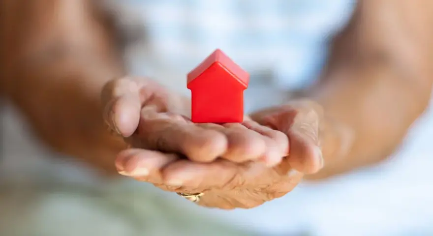 Mains âgées tenant une petite maison rouge. Une image symbolisant la "cosaisine définition" : le foyer, la sécurité, le bien-être.