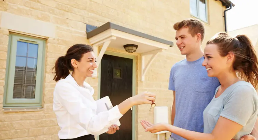 Une femme remet des clés à un jeune couple devant leur nouvelle maison. Le tableau droit de préemption locataire est respecté.