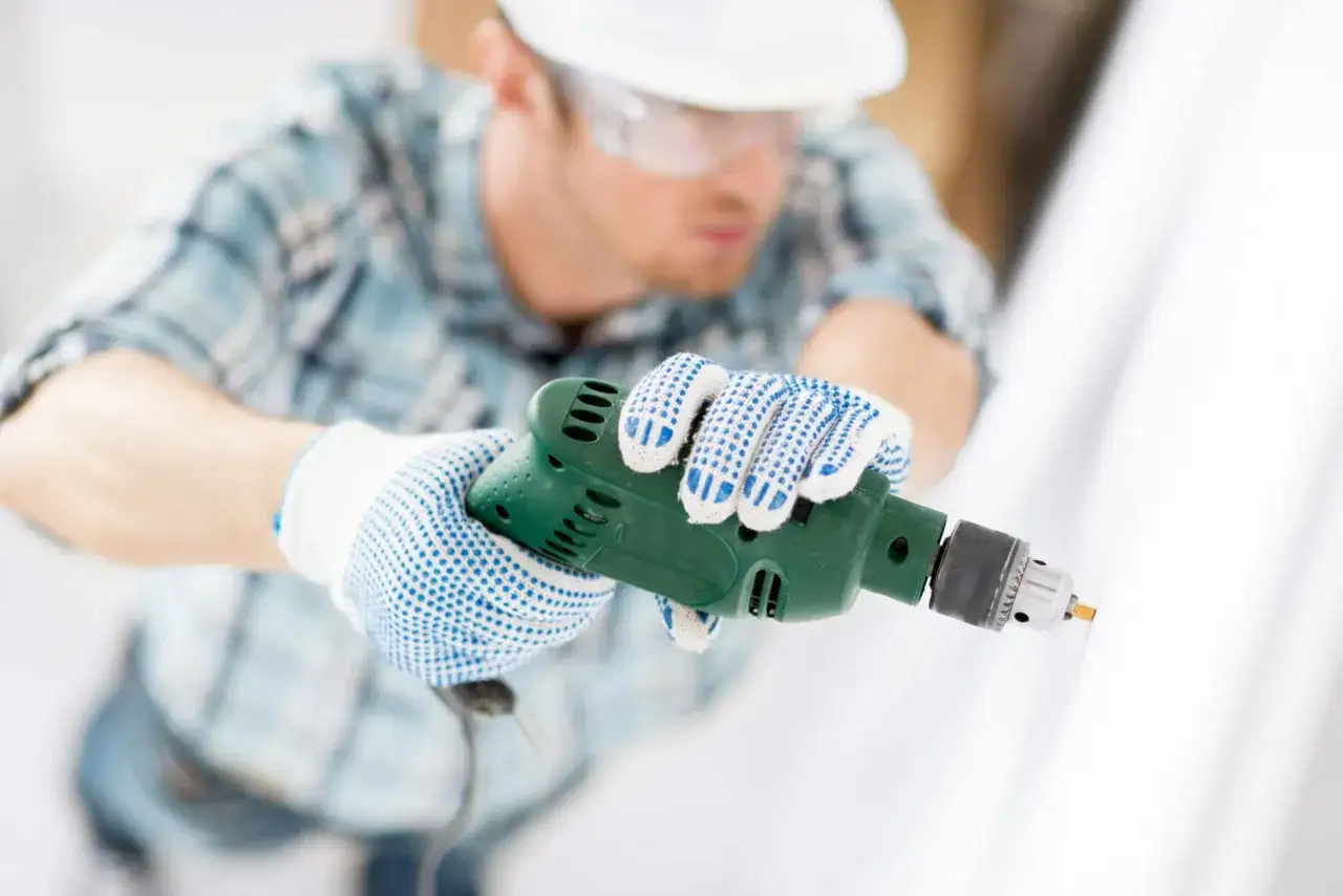Un ouvrier porte des gants bleus et blancs et utilise une perceuse verte pour des travaux d'amélioration.