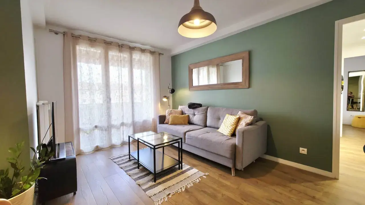 Salon moderne avec canapé gris, table basse en verre, tapis ethnique et mur vert. La t3 définition de ce lieu est un espace cosy et lumineux.
